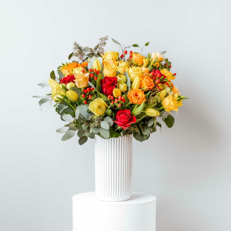 seasonal flowers yellow and orange including roses tulips freesia hypericum berries foliage arranged in a white tall ceramic vase