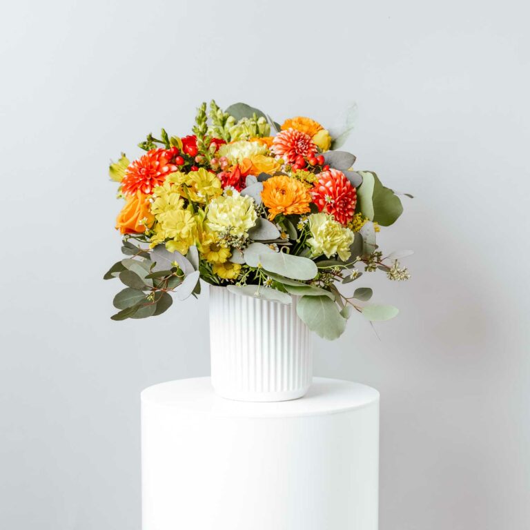seasonal flowers yellow and orange including roses dahlias snapdragons alstromeria calendula carnations chrysanthemums with foliage arranged in a white short ceramic vase