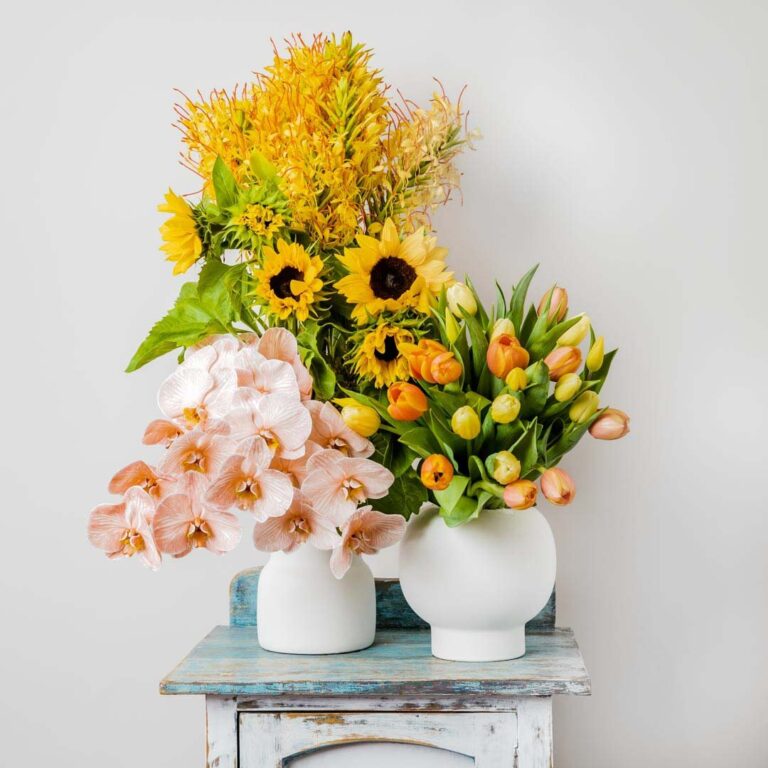 a collection of vibrant yellow and orange flowers arranged in two vases clusters of yellow and orange semi open tulips arranged in a round vase and clusters of long tall sunflowers and ginger flowers creating a display of flowers in cheery bright tones