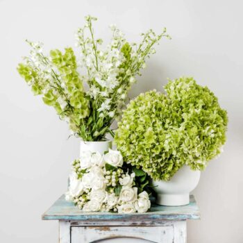a collection of white and green flowers arranged in two vases clusters of green round hydrangea and clusters of long tall white delphinium and lime green irish bells in between a bouquet of white roses with white hypericum berry creating a display of flowers in white and green neutrals