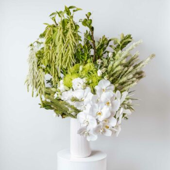 tall vase filled with seasonal white and green flowers including white phalaenopsis orchids lisianthus spider chrysanthemums fig branches woolly bush snowberry and hanging amaranthus