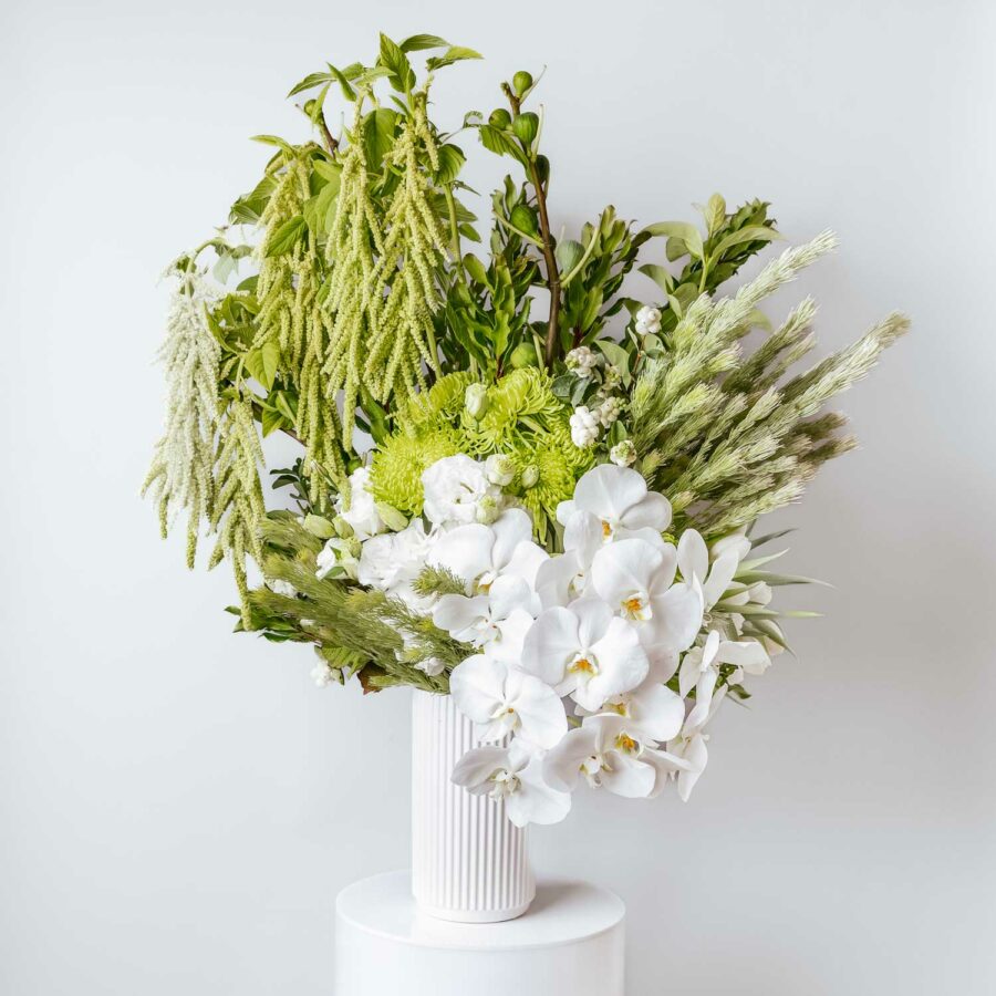 tall vase filled with seasonal white and green flowers including white phalaenopsis orchids lisianthus spider chrysanthemums fig branches woolly bush snowberry and hanging amaranthus