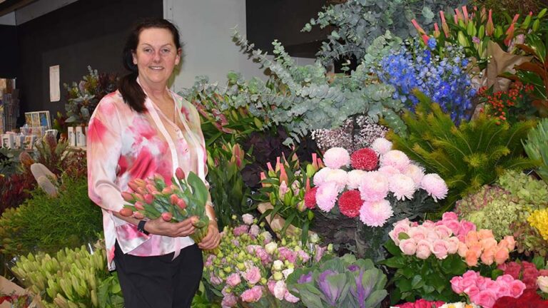 Hermina is standing in front of a vertical floral display of pink, purple and blue flowers as well as blue gum