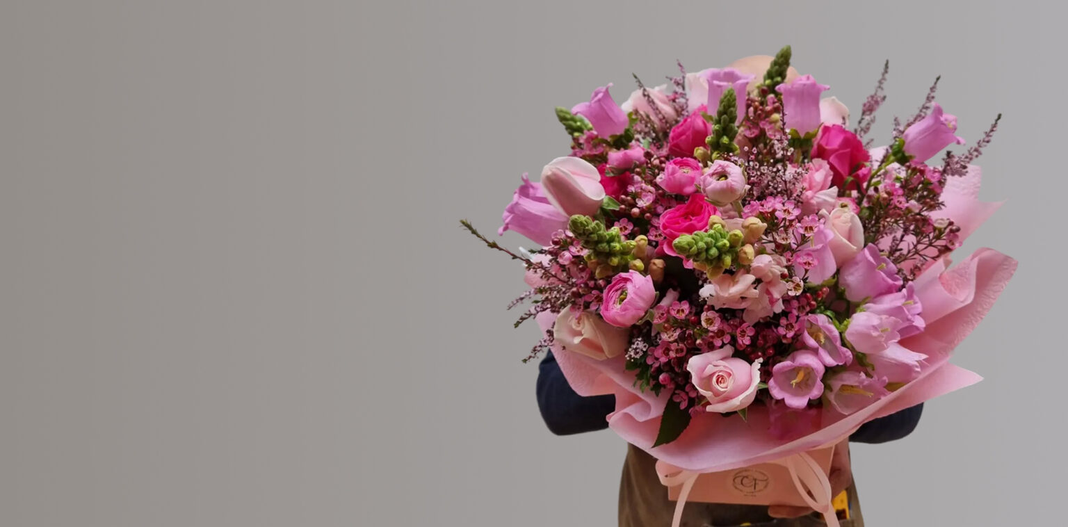 Large bouquet of fresh pink and purple flowers including roses, ranunculus, and Canterbury bells wrapped in soft pink paper